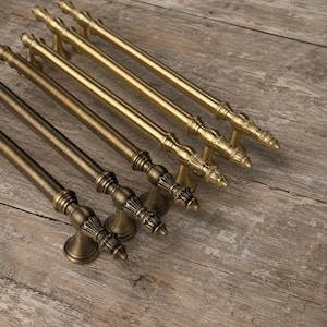 May include: A set of five long, cylindrical metal cabinet pulls arranged in a diagonal row. Each handle features a smooth, straight bar design with decorative, fluted, bell-shaped end caps and circular mounting bases in a brushed brass finish.