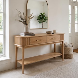 May include: A light oak console table featuring two drawers with thin gold-toned handles, a lower shelf, and rounded sides with vertical fluted detailing. The table surface holds a ceramic vase with dried branches and two stacked books.