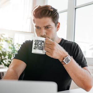 May include: White ceramic mug with the text "ERROR 404: TOLERANCE NOT FOUND" in black. The mug is held by a person wearing a black t-shirt and a watch with a brown leather strap. The person is sitting at a desk.