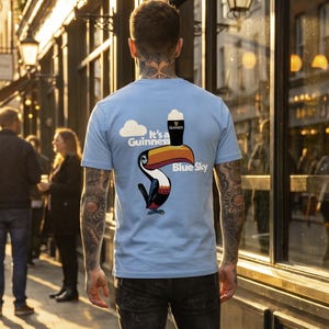 May include: Light blue t-shirt with a Guinness toucan graphic. The design includes a toucan with a pint of Guinness, a cloud, and the words "It's a Guinness Blue Sky". The person is wearing black jeans.