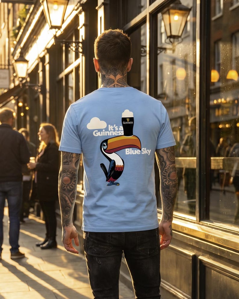 May include: Light blue t-shirt with a Guinness toucan graphic. The design includes a toucan with a pint of Guinness, a cloud, and the words "It's a Guinness Blue Sky". The person is wearing black jeans.