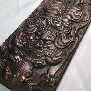 Può includere: Targa decorativa rettangolare color bronzo con un'intagliatura a rilievo dettagliata di una creatura mitica. La creatura ha un'espressione feroce, con dettagli intricati nella criniera, nel corpo e negli elementi decorativi. La targa ha una superficie strutturata.