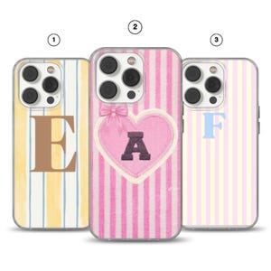 Puede incluir: Tres fundas para smartphone con rayas verticales. La primera tiene rayas amarillas y azules con una letra E marrón grande. La segunda tiene rayas rosas, un corazón central con un lazo rosa y una letra A oscura. La tercera tiene rayas rosas y amarillas con una letra F azul claro.