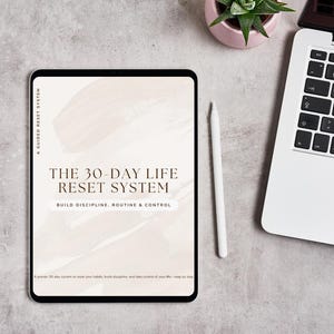 May include: A digital tablet displays "THE 30-DAY LIFE RESET SYSTEM" with the text "BUILD DISCIPLINE, ROUTINE & CONTROL." A white stylus and a laptop are nearby. A small potted plant sits to the side on a gray surface.