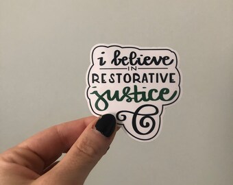May include: A white sticker with the phrase "i believe in restorative justice" in black and green script. The sticker has a cloud-like border and is held by a hand with black nail polish.