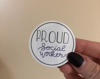 May include: A white, circular sticker with a black border. The sticker reads "PROUD Social Worker" in a mix of bold and cursive fonts. The sticker is held by a hand with dark nail polish.