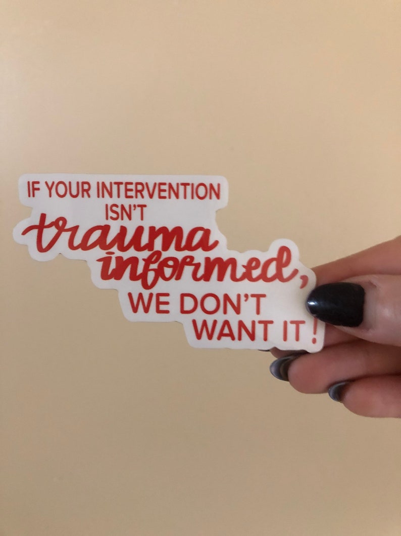May include: A white sticker with red text that reads "IF YOUR INTERVENTION ISN'T trauma informed, WE DON'T WANT IT!" The sticker is held by a hand with dark nail polish against a beige background.