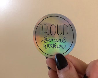 May include: A round, holographic sticker with the words "PROUD Social Worker" in black script. The sticker has a silver border and is held by a hand with black nail polish.