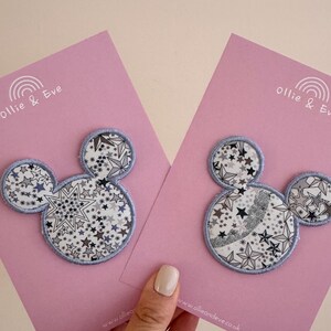 Puede incluir: Dos parches con forma de Mickey Mouse con un estampado de estrellas en blanco, gris y negro, delineados en azul claro. Se muestran en tarjetas rosas con el texto "Ollie & Eve" y una dirección web.