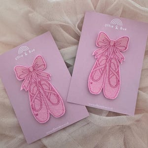 May include: Two pink ballet slipper patches on pink cards. Each patch features a bow and detailed stitching. The cards have the text "Ollie & Eve" and a website address. The background is a soft, textured fabric.