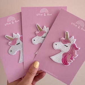 May include: Three embroidered unicorn patches on pink cards. Each patch features a white unicorn head with a gold horn and pink mane. The cards are printed with the text "Ollie & Eve" and a website address. The patches are ideal for clothing.