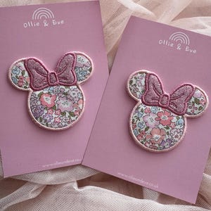 May include: Two Minnie Mouse-shaped fabric patches with floral patterns and glittery pink bows. The patches are on pink cards with the text "Ollie & Eve" and a website address. The background is a soft, light pink fabric.