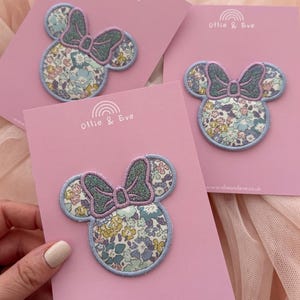 May include: Three Minnie Mouse-shaped patches on pink cards. The patches have a floral pattern in blue, yellow, and white, with a glittery bow and a light blue outline. The cards display the text "Ollie & Eve" and "www.ollieandeve.co.uk".
