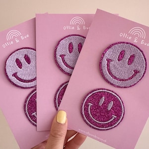 May include: Pink glitter smiley face patches on pink cards. Each patch has a stitched design with a smiling face. The cards have the text "Ollie & Eve" and a rainbow graphic. The website address www.ollieandeve.co.uk is also visible.