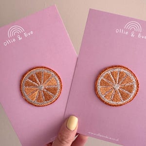 May include: Two embroidered orange slice patches on pink cards. The patches feature orange and white thread, with a silver border. The cards display the text "Ollie & Eve" and a rainbow graphic. The website address www.ollieandeve.co.uk is also visible.