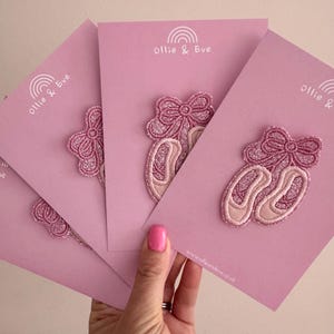 May include: Pink ballet slipper patches with glitter bows on pink cards. The cards feature the text "Ollie & Eve" and a rainbow logo. The website address www.ollieandeve.co.uk is printed on the cards.