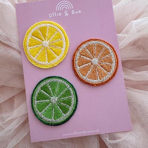 May include: Embroidered patches featuring citrus fruit slices: a yellow lemon, an orange, and a green lime. The patches are on a pink card with the text "Ollie & Eve" and a rainbow graphic. The website address www.ollieandeve.co.uk is also visible.