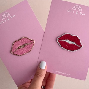 May include: Two embroidered lip-shaped patches on light pink backing cards. One patch is light pink with a gold glitter border, and the other is dark red with a silver glitter border. Each card features the text Ollie & Eve with a rainbow icon.
