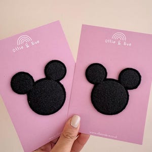 Könnte beinhalten: Zwei schwarze Filz-Mickey-Mouse-Kopf-Patches auf rosa Karten. Die Karten tragen den Text "Ollie & Eve" und ein Regenbogen-Logo. Die Website-Adresse www.ollieandeve.co.uk ist unten aufgedruckt. Die Patches werden von einer Person gehalten.