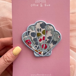 May include: A fabric seashell-shaped patch with a floral design in blue, green, and red, edged with light blue stitching. The patch is on a pink card with the text "ollie & Eve" and a rainbow logo.