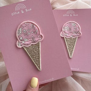 Puede incluir: Dos tarjetas rosas con parches de cono de helado. Las bolas de helado tienen un estampado floral y los conos son brillantes. El texto "Ollie & Eve" y un arcoíris están impresos en las tarjetas. La dirección del sitio web ollieandeve.co.uk está impresa en la parte inferior de las tarjetas.