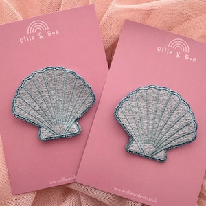 May include: Two embroidered seashell patches with light blue thread and glitter trim are displayed on pink cards. The cards have the text "Ollie & Eve" and a rainbow logo. The background is a soft, pink fabric.