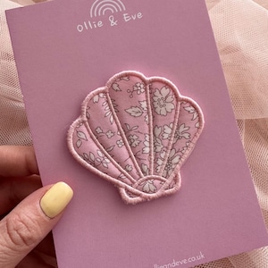 May include: A pink seashell-shaped fabric patch with a floral design, attached to a pink card. The patch has a light pink outline and a pattern of white flowers and leaves. The card displays the text "Ollie & Eve" and "www.ollieandeve.co.uk".