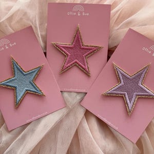 May include: Three star-shaped embroidered patches in pastel shades of blue, pink, and purple. Each star has a gold glitter border and is mounted on a pink card. The cards display the text "Ollie & Eve" and a website address.