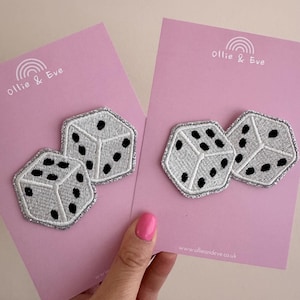 May include: Two dice patches with a silver border and black dots are displayed on pink cards. The patches are embroidered with a white thread. The cards have the text "Ollie & Eve" and a rainbow design.