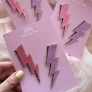 May include: Embroidered lightning bolt patches in pink and purple with gold glitter trim, presented on pink cards. The cards feature the text "Ollie & Eve" and a website address. A playful accessory.
