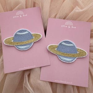 May include: Two embroidered planet patches. Each patch features a light blue planet with a gold ring, set against a pink card. The cards have the text "Ollie & Eve" and "www.ollieandeve.co.uk".