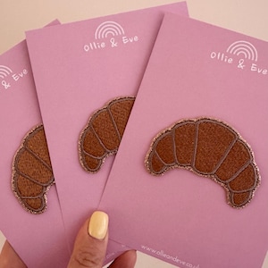 May include: Three embroidered croissant patches on pink cards. The patches are brown with gold glitter trim. The cards have the text "ollie & Eve" and a rainbow graphic. The website address www.ollieandeve.co.uk is also visible.