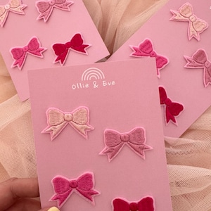 May include: Pink cards featuring embroidered bow patches in shades of pink and red. The cards display the text "Ollie & Eve" and a rainbow logo. The website address www.ollieandeve.co.uk is printed at the bottom of the cards.