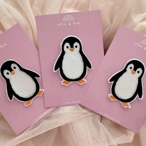 Könnte beinhalten: Drei gestickte Pinguin-Aufnäher auf rosa Karten. Jeder Pinguin hat einen schwarzen Körper, einen weißen Bauch, orangefarbene Füße und einen weißen Schnabel. Die Karten tragen den Text "Ollie & Eve" und ein Regenbogen-Logo.