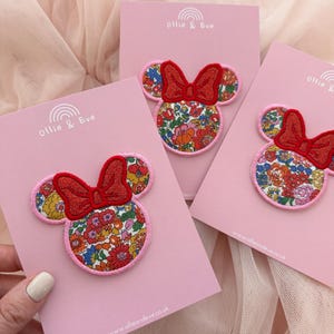 Puede incluir: Tres parches con forma de Minnie Mouse con lazos rojos y caras con estampado floral se muestran en tarjetas rosas. Los parches están sobre un fondo rosa con el nombre de la marca "Ollie & Eve" y la dirección del sitio web.