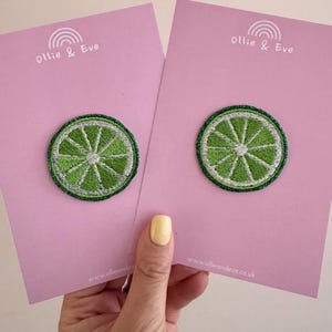 May include: Two embroidered lime slice patches on pink cards. The patches feature a green lime design with white details and a white border. The cards have the text "Ollie & Eve" and a website address.