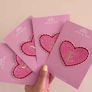May include: A hand holding several pink cards with embroidered heart patches. Each heart patch has a gold letter and a red scalloped border. The cards have the text "Ollie & Eve" and a rainbow design.