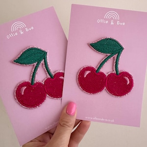 May include: Two cherry patches with red cherries, green leaves, and glittery pink borders are displayed on pink cards. The cards have the text "Ollie & Eve" and a rainbow logo. The website address www.ollieandeve.co.uk is also visible.