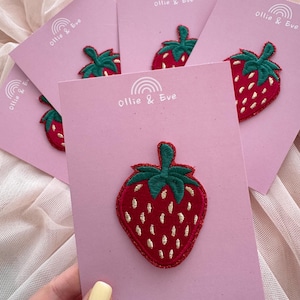 May include: A collection of strawberry patches on pink cards. The patches are red with white seed details and green stems. The cards are branded with "Ollie & Eve" and the website address.