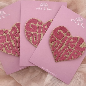 May include: Three pink cards with heart-shaped patches that read "GIRL POWER" in gold glitter lettering. The patches are embroidered and have a pink background. The cards have the brand name "Ollie & Eve" and a rainbow logo.