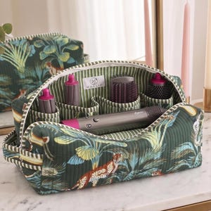 May include: A quilted fabric storage bag featuring a dark green jungle print with cheetahs and foliage. The interior is lined with green and white stripes and contains a grey hair styling tool with magenta accents and several matching attachments.