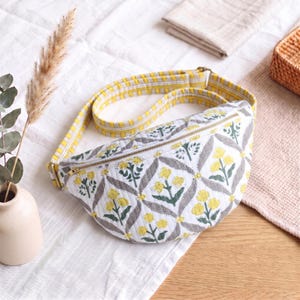 May include: A quilted fanny pack featuring a white base with a repeating gray diamond pattern and yellow floral motifs. The bag includes a gold-tone zipper closure and an adjustable strap with a yellow and white vertical striped fabric design.