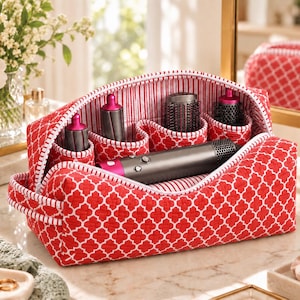 May include: A red and white quilted fabric storage case with a geometric pattern, featuring an open top and interior compartments holding a gray and fuchsia hair styling tool and several cylindrical attachments in matching colors.