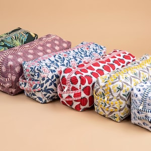 K&ouml;nnte beinhalten: Sechs rechteckige, gesteppte Stofftaschen in einer Reihe. Jede Tasche hat ein anderes Muster, darunter dunkelgr&uuml;nes Blattwerk, violette Blumen, blaue Blumen, rote Erdbeeren, gelbe Blumen und blaue geometrische Drucke mit Rei&szlig;verschl&uuml;ssen.