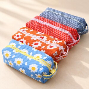 May include: Four rectangular zippered pouches arranged in a row. The front pouch is blue with white and yellow daisies, followed by an orange pouch with white daisies, a red pouch with a white geometric pattern, and a blue geometric pouch.