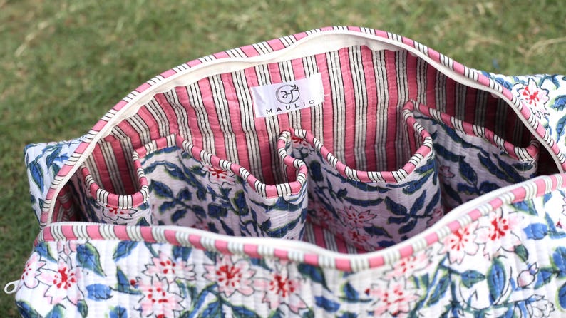 K&ouml;nnte beinhalten: Eine gesteppte Stofftasche mit Rei&szlig;verschluss, die au&szlig;en wei&szlig; mit einem blau-rosa Blumenmuster bedruckt ist. Das Innere ist mit einem rosa-wei&szlig; gestreiften Stoff gef&uuml;ttert und verf&uuml;gt &uuml;ber mehrere offene Taschen. Ein wei&szlig;es Etikett tr&auml;gt die Aufschrift MAULIO.