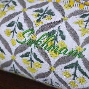K&ouml;nnte beinhalten: Ein wei&szlig;er, gesteppter Stoffbeutel mit einem sich wiederholenden Muster aus gelben Blumen und grauen Bl&auml;ttern, wobei der Name Sukhmani in gr&uuml;ner Schreibschrift in der Mitte eingestickt ist und eine gelb-wei&szlig; gestreifte Kante oben.