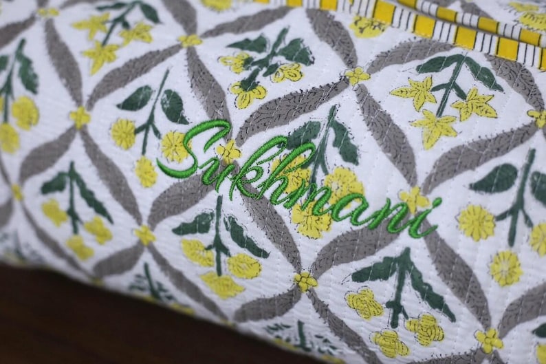 K&ouml;nnte beinhalten: Ein wei&szlig;er, gesteppter Stoffbeutel mit einem sich wiederholenden Muster aus gelben Blumen und grauen Bl&auml;ttern, wobei der Name Sukhmani in gr&uuml;ner Schreibschrift in der Mitte eingestickt ist und eine gelb-wei&szlig; gestreifte Kante oben.