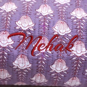 K&ouml;nnte beinhalten: Ein gesteppter Stoff mit einem sich wiederholenden Muster aus wei&szlig;en glockenf&ouml;rmigen Bl&uuml;ten mit roten Umrissen auf hellviolettem Grund. Der Name Mehak ist in roter Schreibschrift in der Mitte der gesteppten Textiloberfl&auml;che eingestickt.