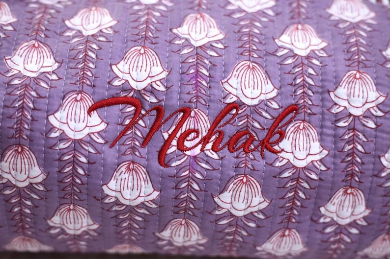 K&ouml;nnte beinhalten: Ein gesteppter Stoff mit einem sich wiederholenden Muster aus wei&szlig;en glockenf&ouml;rmigen Bl&uuml;ten mit roten Umrissen auf hellviolettem Grund. Der Name Mehak ist in roter Schreibschrift in der Mitte der gesteppten Textiloberfl&auml;che eingestickt.